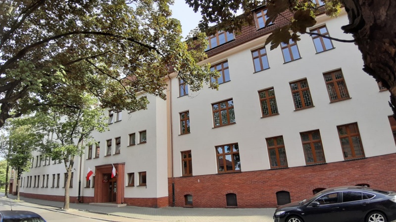 Widok fasady budynku urzędu z oknami i drzwiami wejściowymi. Przed bydynkiem rosnace drzewa.Obok stojący samochód. 