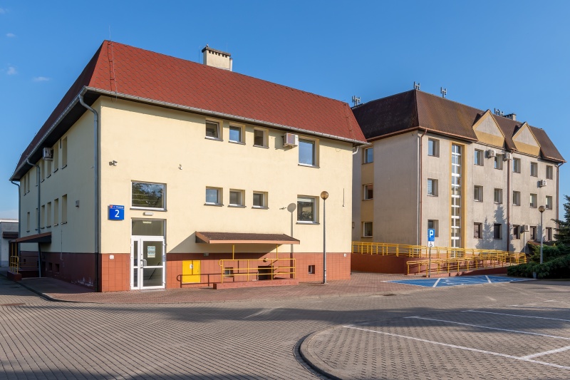 Widok fasady budynku urzędu z oknami. Przed wejsciem podjazd oraz parking z wyznaczonym miejscem dla osób niepełnosprawnych