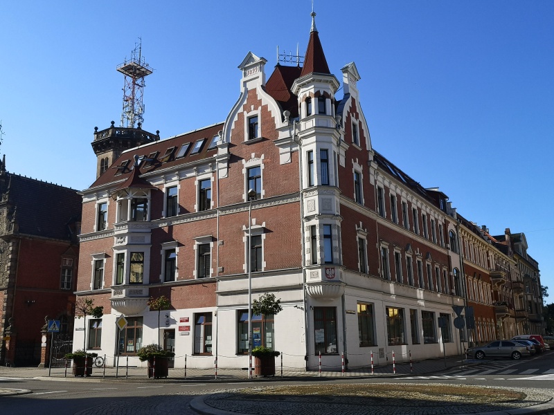 Widok zabytkowej fasady budynku urzędu.Front z cegły, zdobienia nad oknami i ozdobne wieżyczki.Z boku budynku na parkingu stojace samochody.