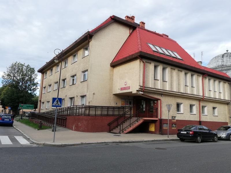 Widok budynku urzędu z oknami i wejściem z podjazdem dla osób niepełnosprawnych.Przed budynkiem na ulicy stoją samochody osobowe.