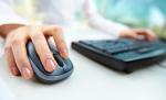 Prawa dłoń kobiety w białej bluzie na myszce do komputera, podczas gdy lewa obsługuje klawiaturę.