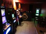 w ciemny pomieszczeniu stoi 6 automatów do gier hazardowych, 5 pod lewą ścianą pomieszczenia, jeden w prawym kącie, wszystkie są włączone, świecą kolorowymi światłami