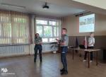 dwóch funkcjonariuszy Służby Celno-Skarbowej stoi przed uczniami, na ekranie na ścianie wyświetlana jest prezentacja