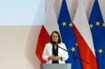 Minister Finansów stoi przy mównicy, w tle flagi RP i UE