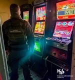 Widać funkcjonariusza Służby Celno-Skarbowej, który stoi przy automatach do gier. W pomieszczeniu jest ciemno i widać jasne i kolorowe ekrany automatów.