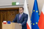 Szef Krajowej Administracji Skarbowej Marcin Łoboda podczas konferencji prasowej przy mównicy w tle flagi Polski i Unii Europejskiej