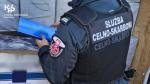 funkcjonariusz Służby Celno-Skarbowej stroi tyłem przed pojazdem wypełnionym workami jutowymi. W dłoniach trzyma karton nielegalnych papierosów