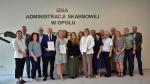 grupa kobiet i mężczyzn, naczelników urzędów skarbowych województwa opolskiego i dyrekcja Izby Administracji Skarbowej w Opolu stoją pod ścianą z napisem Izba Administracji Skarbowej w Opolu. Trzy osoby dwie kobiety i jeden mężczyzna trzymają w dłoniach dyplomy