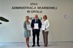  Trzy osoby: dwie kobiety i jeden mężczyzna trzymają w dłoniach dyplomy, stoją pod ścianą z napisem Izba Administracji Skarbowej w Opolu
