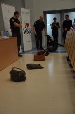 Dwóch funkcjonariuszy opolskiej KAS przygotowuje czarnego psa rasy labrador o imieniu Bella do pokazu polegającego na przeszukaniu leżących na podłodze toreb i plecaka na obecność tytoniu.