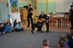 Funkcjonariusze opolskiej KAS w czarnych mundurach służbowych wraz z psem. Funkcjonariusz siedzący na krześle opowiada dzieciom o specyfice pracy psa dziecom.